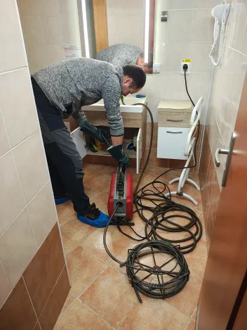 Technik čistí strojem odpad umyvadla v koupelně, hadice smotaná na dlažbě
